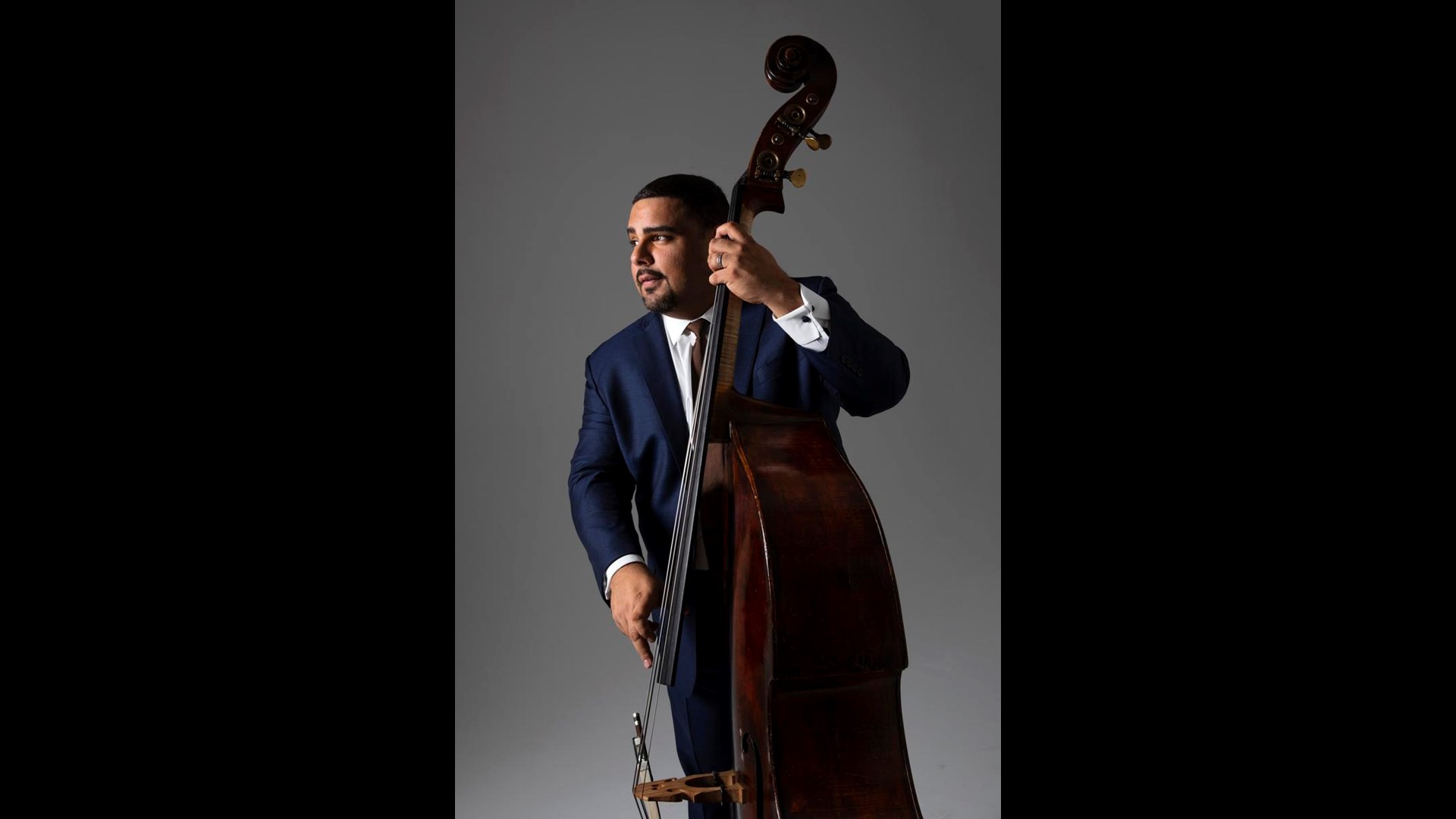 A man with medium complexion and short dark hair in a blue suit playing a double bass against a grey background.