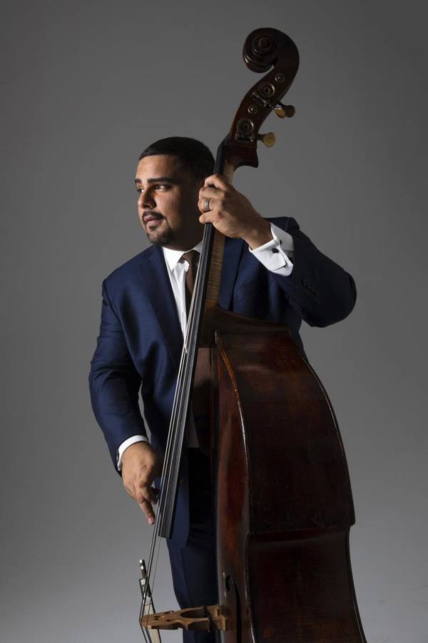 A man with medium complexion and short dark hair in a blue suit playing a double bass against a grey background.