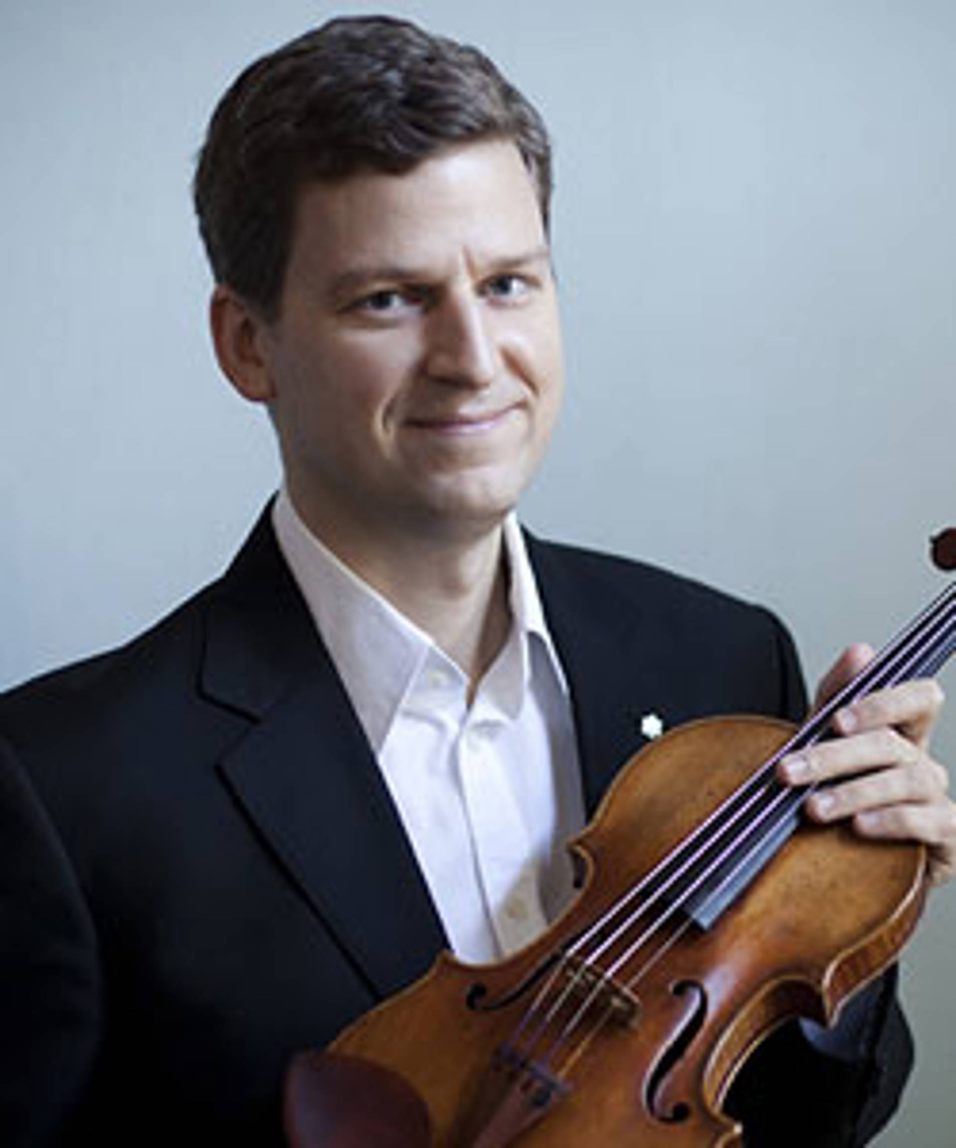 Headshot of a caucasian man holding a violin and smiling. Hey has short brown hair and is wearing a dresshirt and jacket